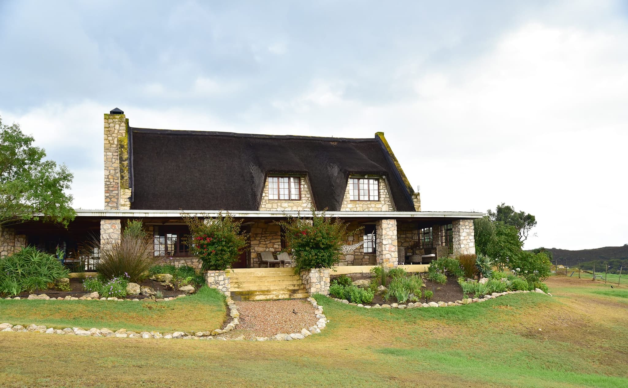 Stonehouse Lodge from the lawn: a thatched stone house with dormer windows, a long covered veranda and a garden path to the steps.